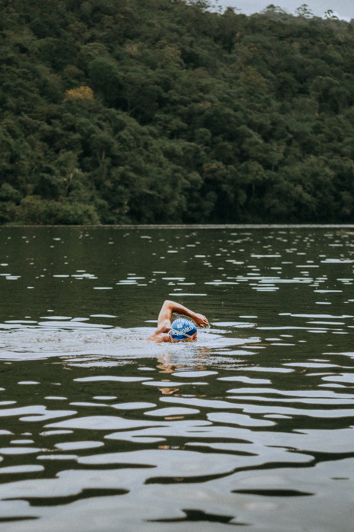 An adult swimmer wearing a blue swim cap trains in a serene forest lake.